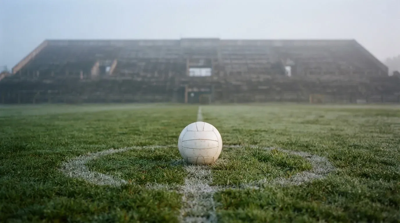 Ein Fußball liegt auf dem Mittelkreis eines leeren Rasenplatzes am frühen Morgen, im Hintergrund eine ferne Tribüne im Dunst.