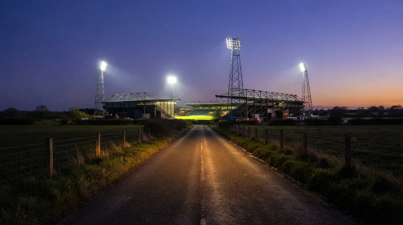Ein Flutlichtstadion erhebt sich bei Dämmerung in der Ferne hinter einer leeren Landstraße, die Lichter sind eingeschaltet.