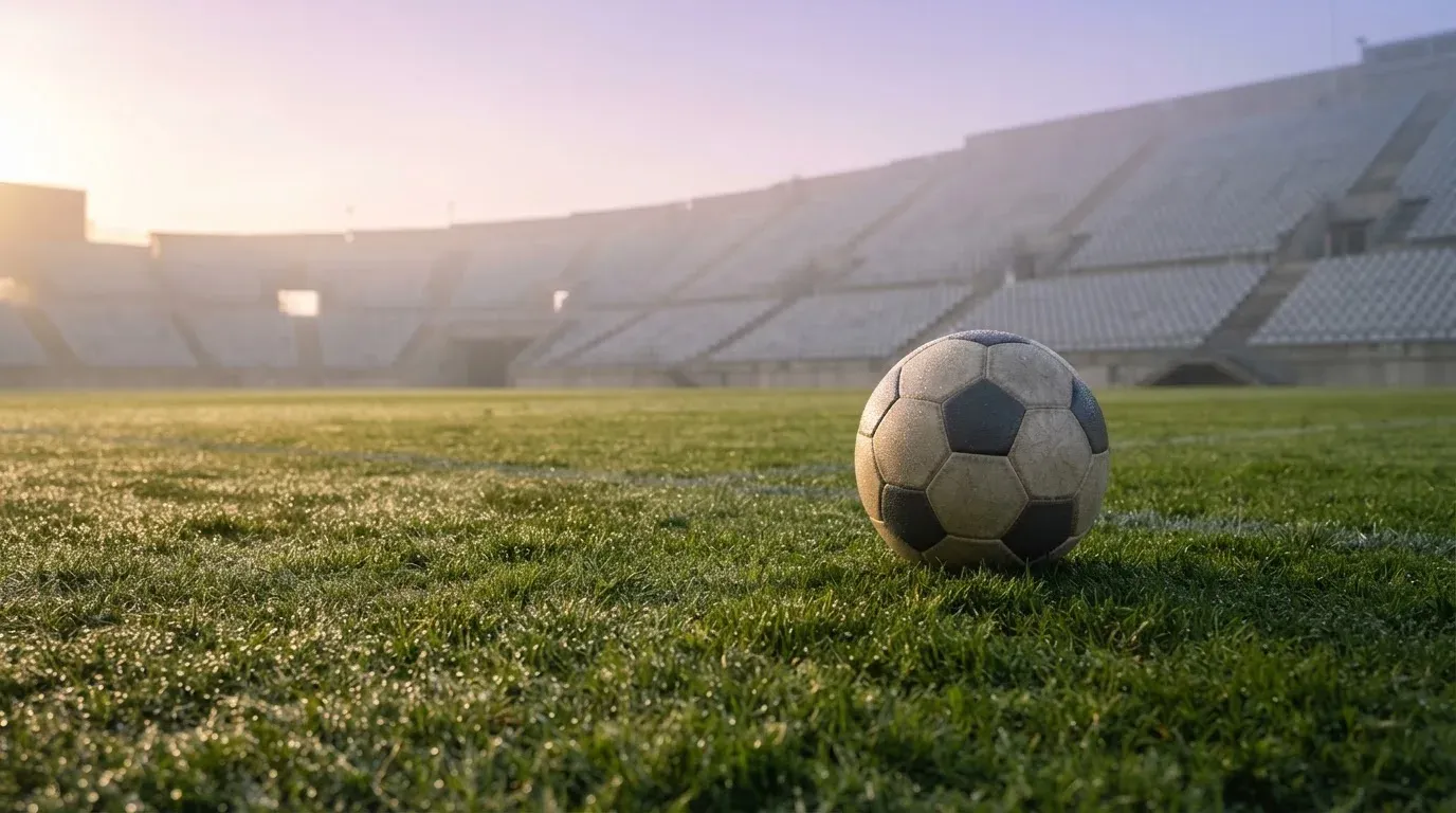 Ein Fußball liegt auf dem Rasen vor einer leeren Tribüne am frühen Morgen, Tau glänzt auf dem Gras.