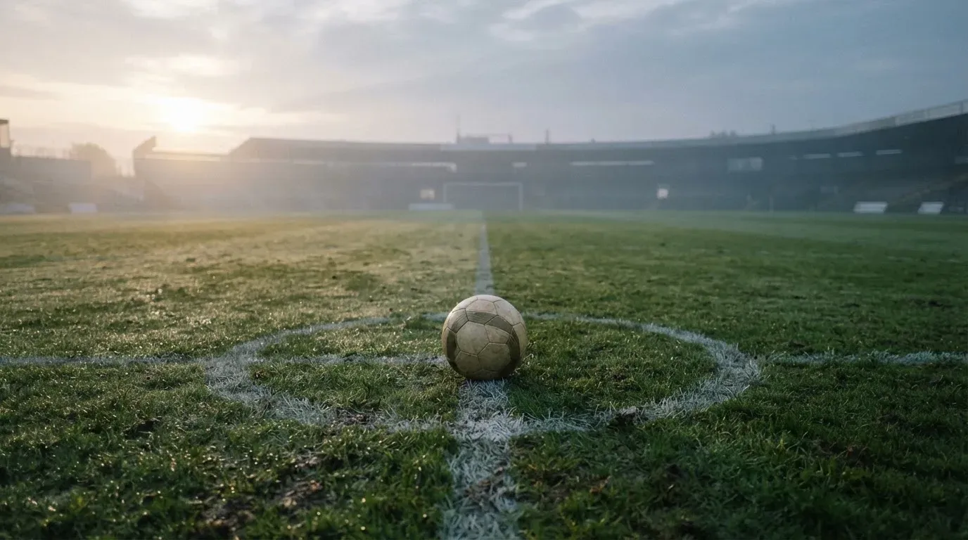 Ein Fußball liegt auf dem Mittelkreis eines leeren Rasenplatzes am frühen Morgen, im Hintergrund eine ferne Tribüne im Dunst.