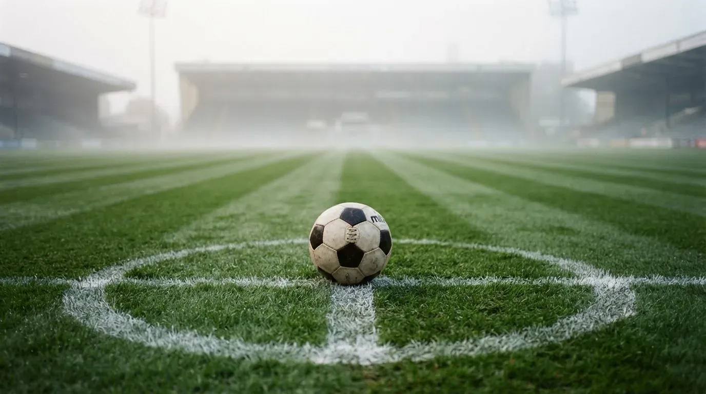 Ein Fußball liegt auf dem Anstoßpunkt eines leeren Rasenplatzes im Morgendunst, im Hintergrund eine ferne Tribüne.