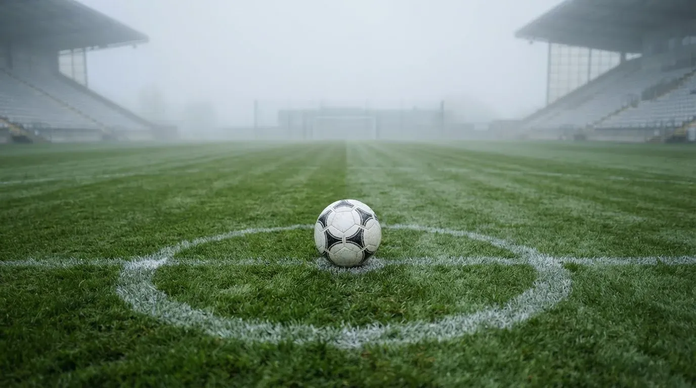 Ein Fußball liegt auf dem Anstoßpunkt eines leeren Rasenplatzes im Morgennebel, im Hintergrund verschwimmen die Tribünen.