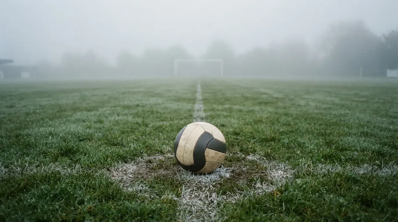 Ein Fußball liegt auf dem Anstoßpunkt eines leeren Rasenplatzes im Morgennebel.