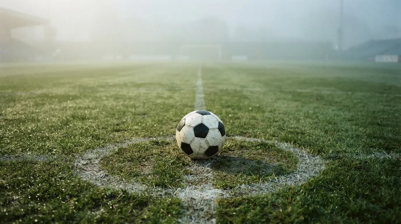 Ein Fußball liegt auf dem Anstoßpunkt eines leeren Rasenplatzes im Morgennebel.