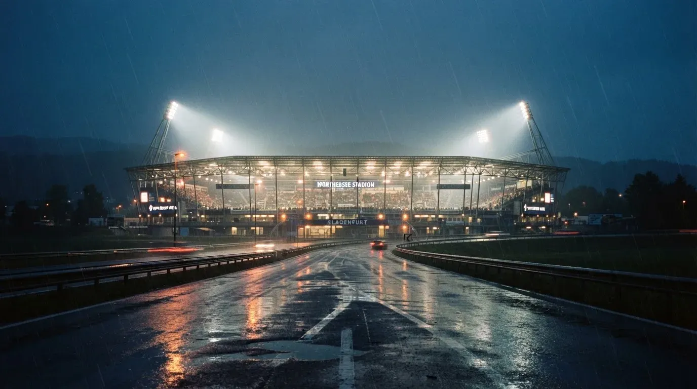 Ein Stadion unter Flutlicht aus großer Entfernung gesehen, davor eine regennasse Straße in der Dämmerung.