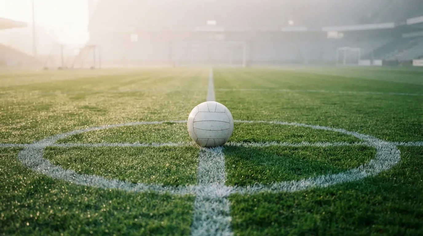 Ein Fußball liegt auf dem Anstoßpunkt eines leeren Rasenplatzes im Morgennebel.