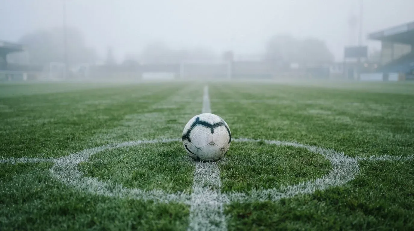 Ein Fußball liegt auf dem Anstoßpunkt eines leeren Rasenplatzes im Morgennebel.