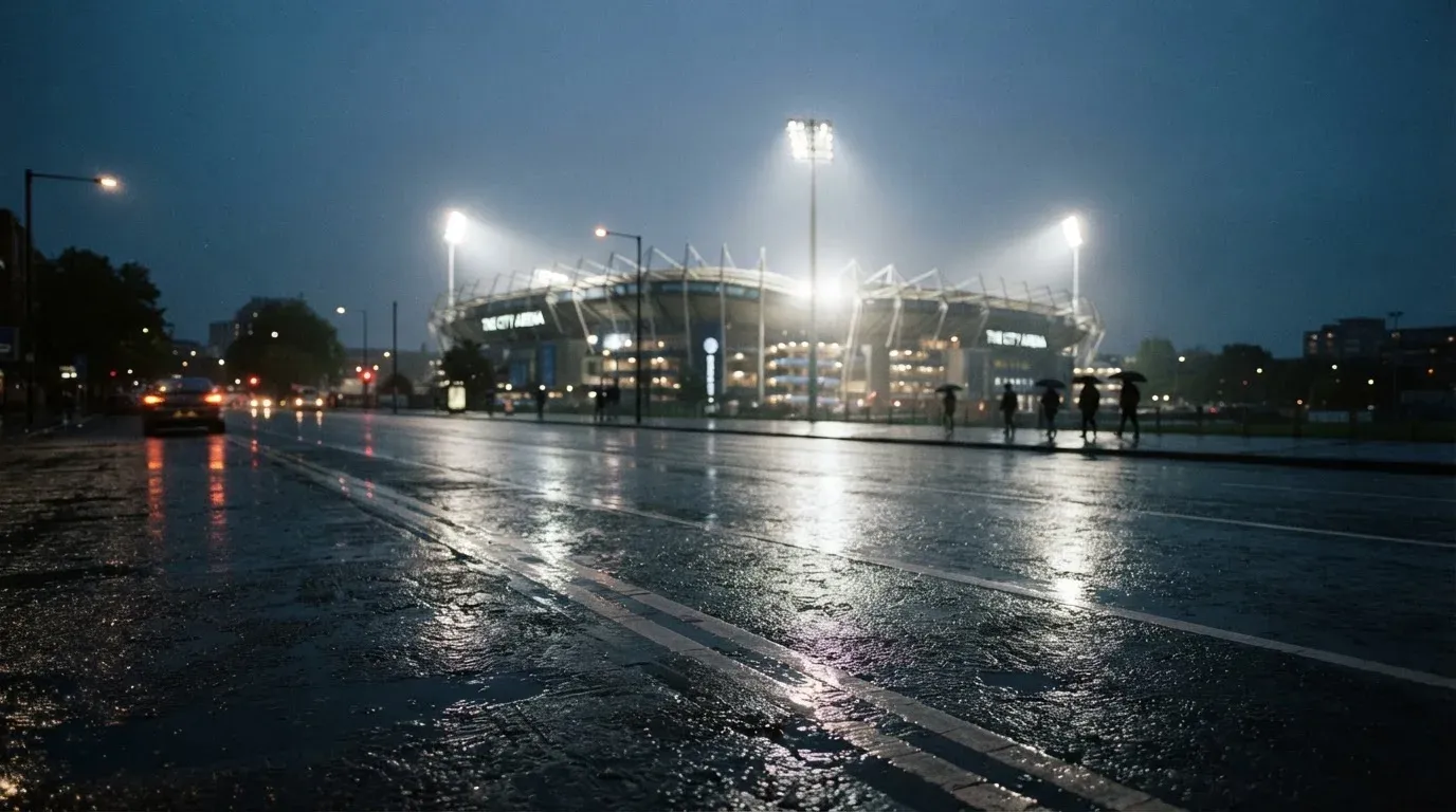 Ein Stadion unter Flutlicht aus großer Entfernung gesehen, davor eine regennasse Straße in der Dämmerung.