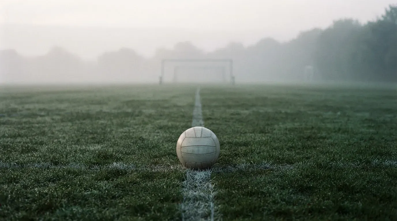 Ein Fußball liegt auf dem Anstoßpunkt eines leeren Rasenplatzes im Morgennebel.