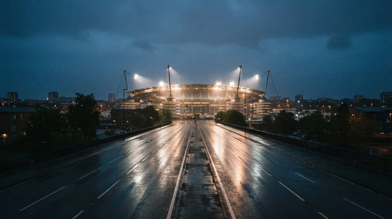 Ein Stadion unter Flutlicht aus großer Entfernung gesehen, davor eine regennasse Straße in der Dämmerung.