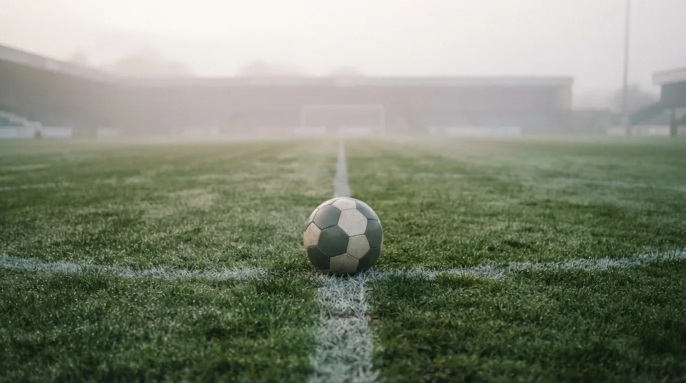 Ein Fußball liegt auf dem Anstoßpunkt eines leeren Rasenplatzes im Morgennebel.