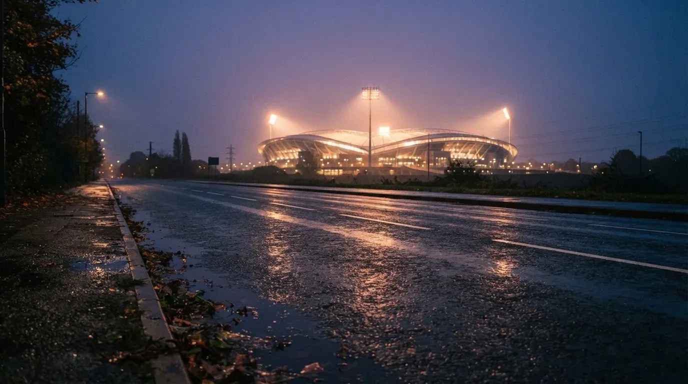 Ein Stadion unter Flutlicht aus großer Entfernung gesehen, davor eine regennasse Straße in der Dämmerung.