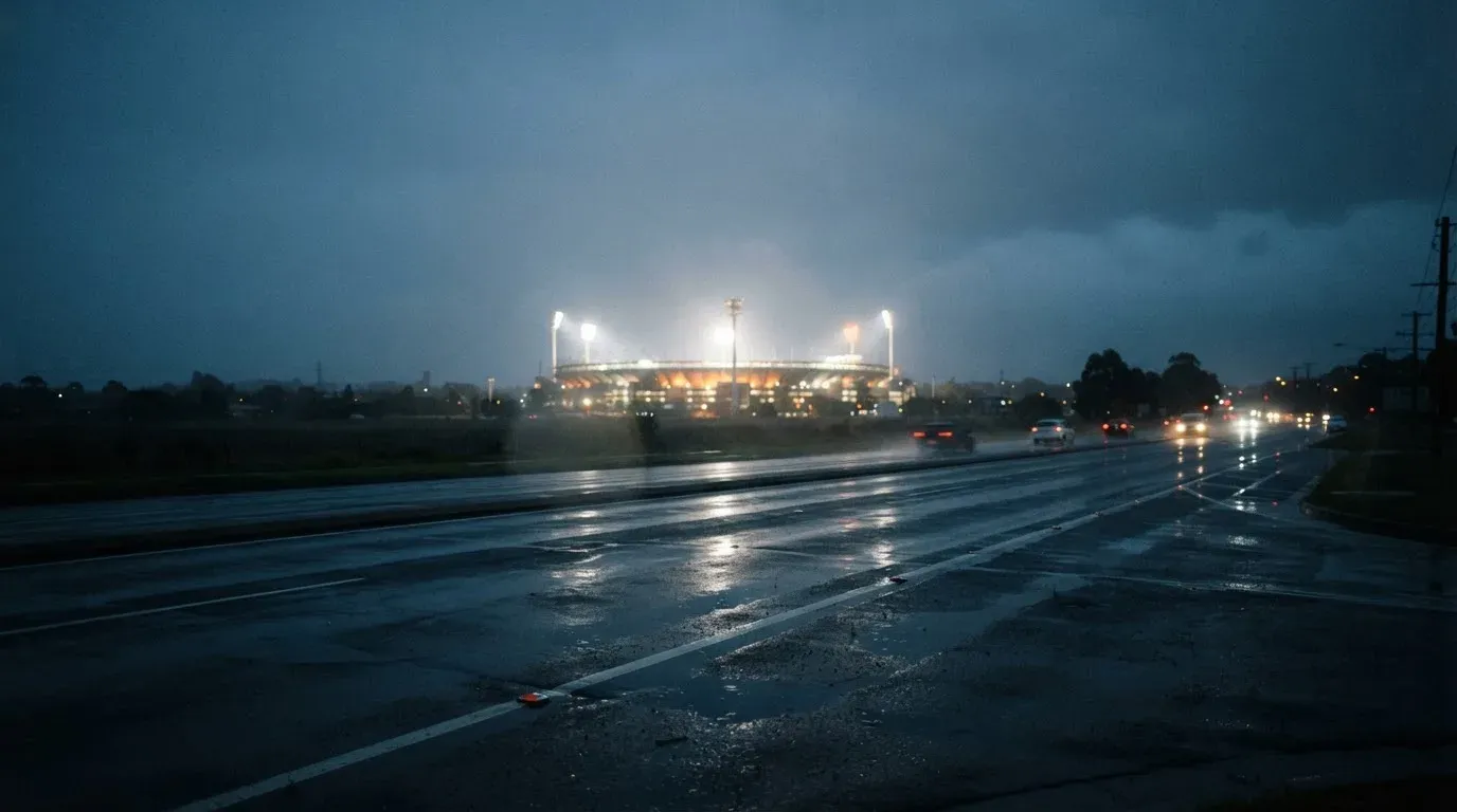 Ein Stadion unter Flutlicht aus großer Entfernung gesehen, davor eine regennasse Straße in der Dämmerung.