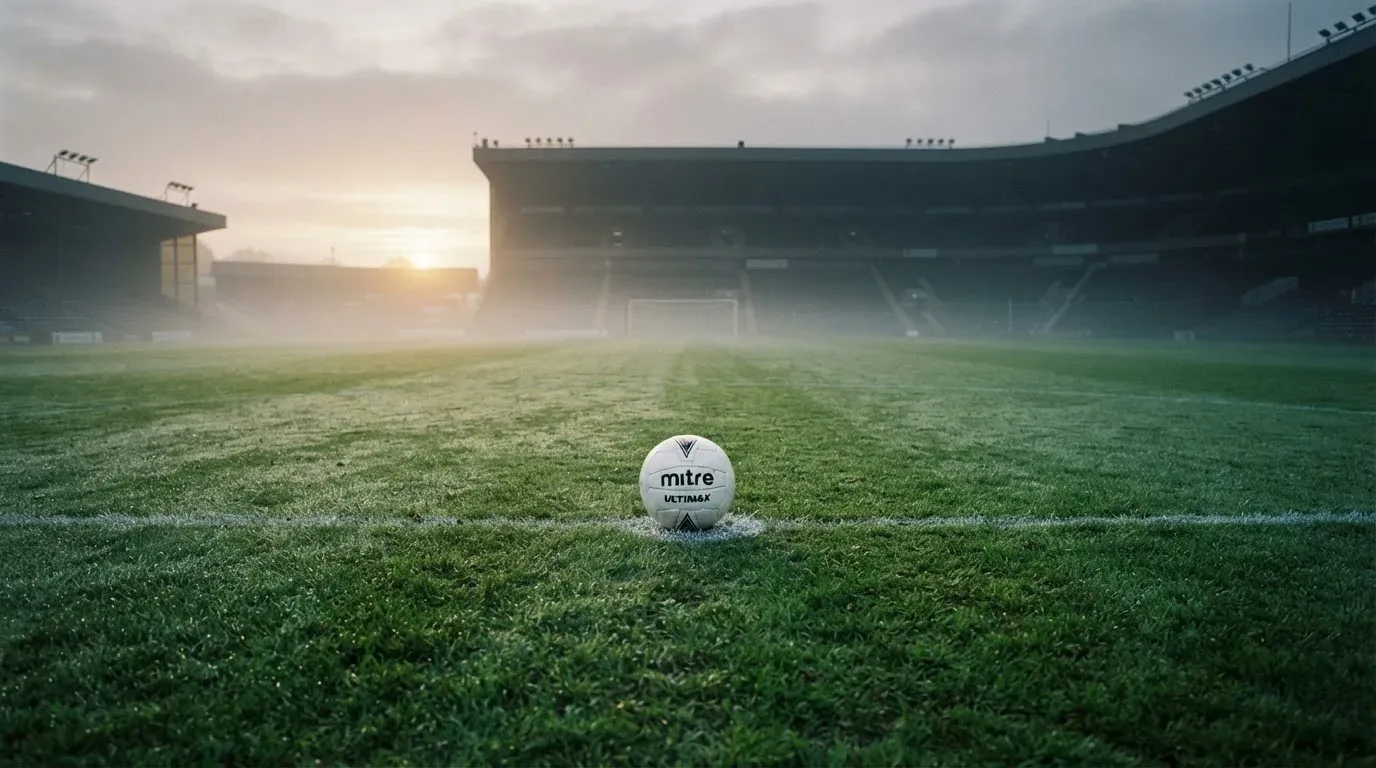 Ein Fußball liegt auf dem Anstoßpunkt eines leeren Rasenplatzes im Morgennebel.