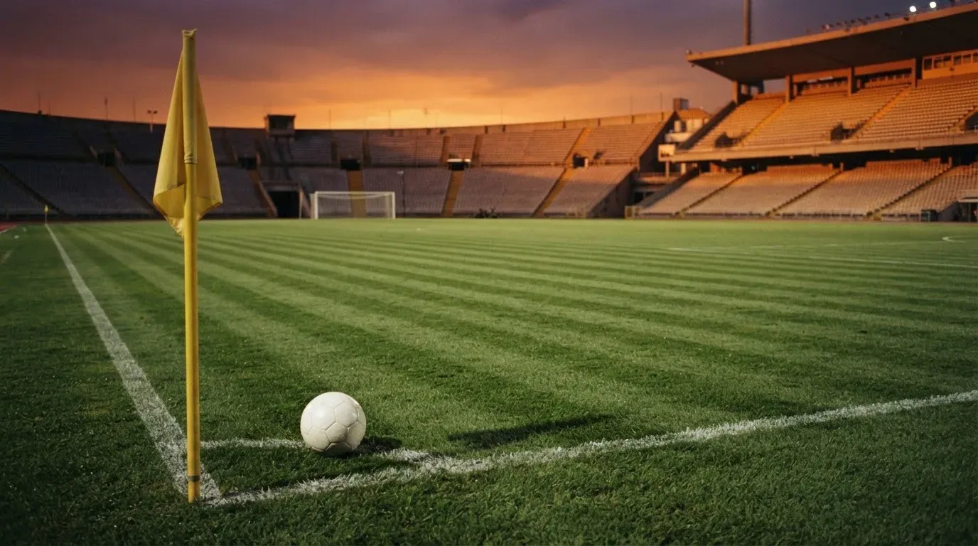 Ein Fußball liegt auf dem Rasen neben der Eckfahne eines leeren Platzes, dahinter eine Tribüne im Abendlicht.