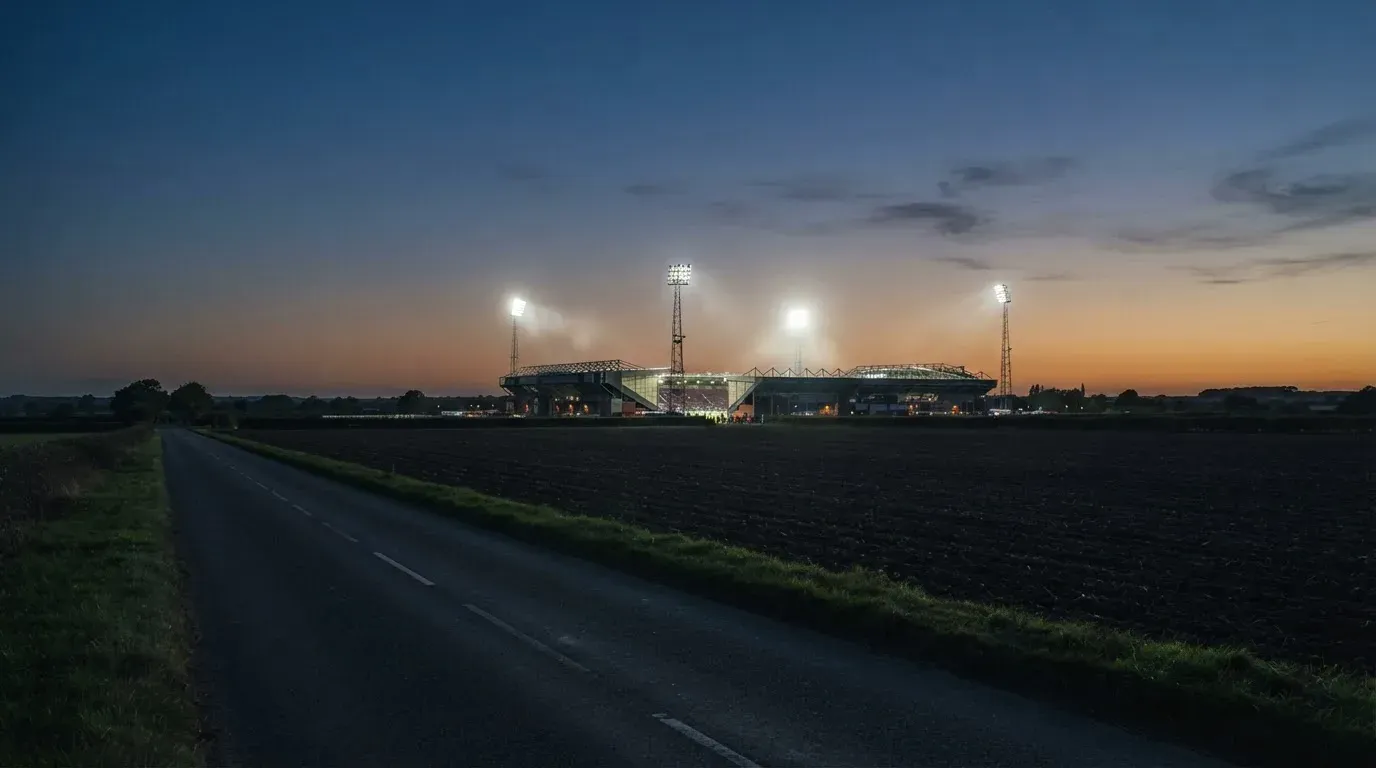 Ein Stadion unter Flutlicht aus großer Entfernung, davor Felder und eine ruhige Straße in der Dämmerung.