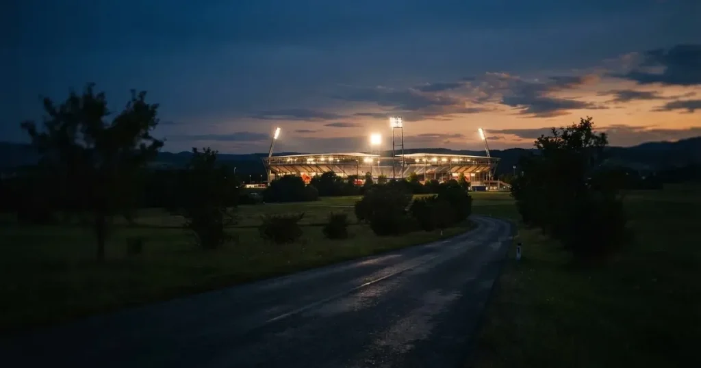 Ein Stadion unter Flutlicht aus großer Entfernung gesehen, davor eine dunkle Landstraße und einzelne Bäume in der Abenddämmerung.