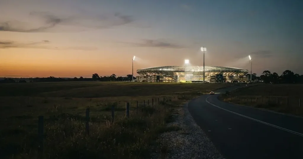 Ein Fußballstadion unter Flutlicht aus großer Entfernung gesehen, davor Felder und eine ruhige Straße in der Dämmerung.