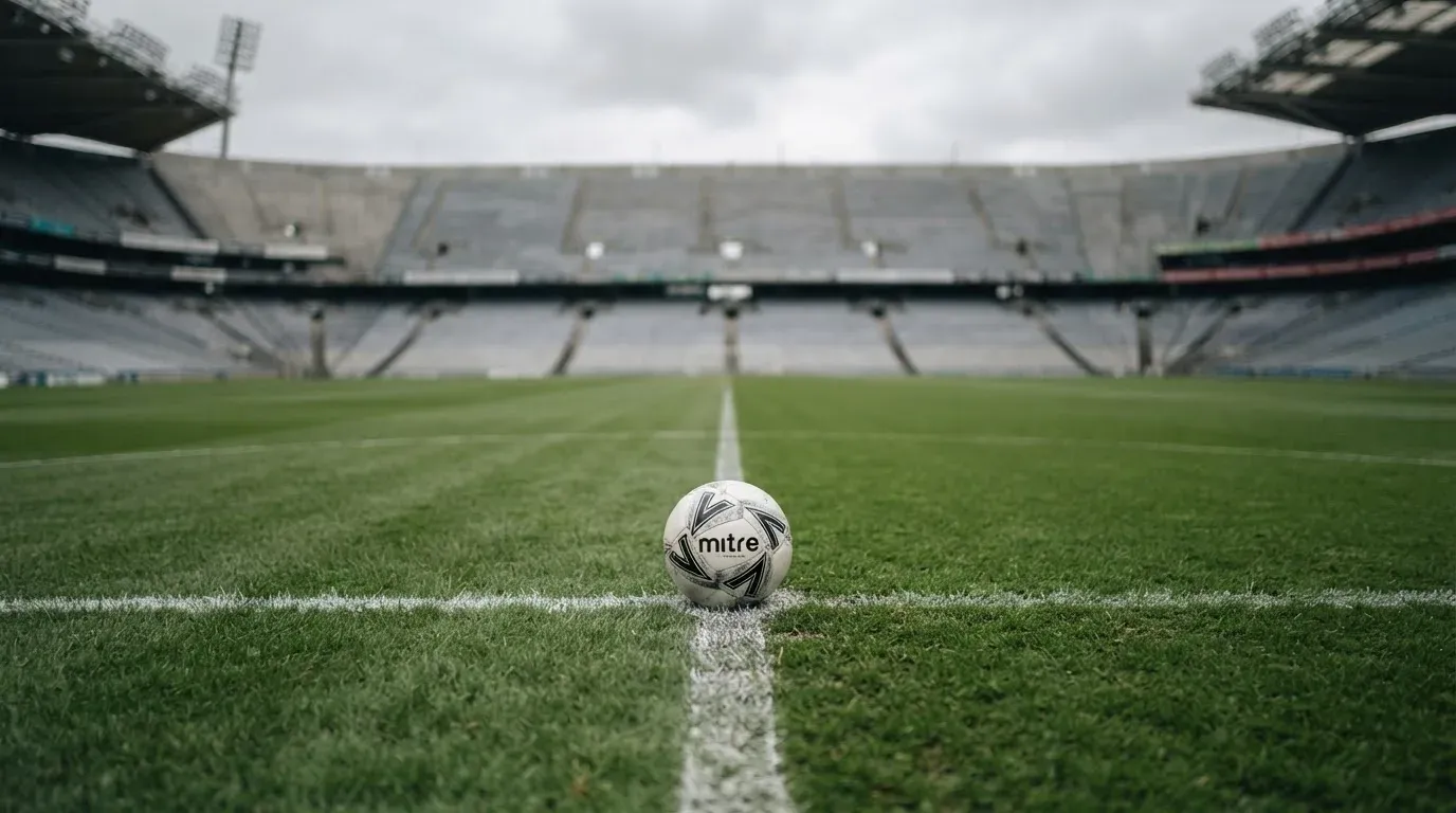 Ein Fußball liegt auf dem Anstoßpunkt eines leeren Rasenplatzes, dahinter eine leere Tribüne in weichem Licht.