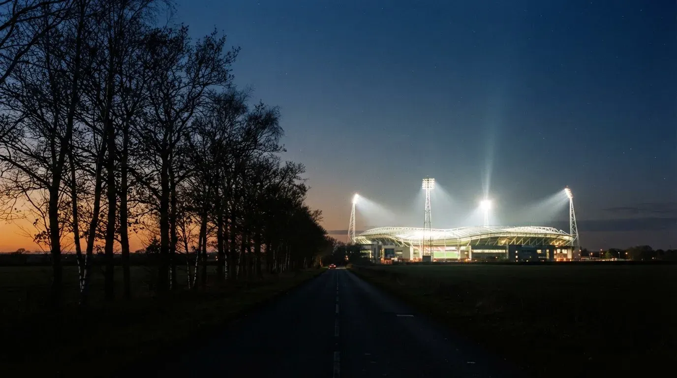 Ein Stadion unter Flutlicht aus großer Entfernung gesehen, davor eine dunkle Landstraße und einzelne Bäume in der Dämmerung.