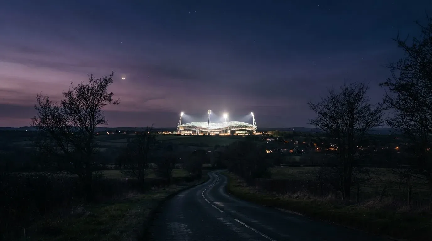 Ein Stadion unter Flutlicht aus großer Entfernung gesehen, davor eine dunkle Landstraße und einzelne Bäume in der Dämmerung.