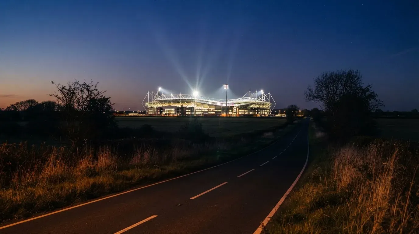 Ein Stadion unter Flutlicht aus großer Entfernung gesehen, davor eine dunkle Landstraße und einzelne Bäume in der Dämmerung.