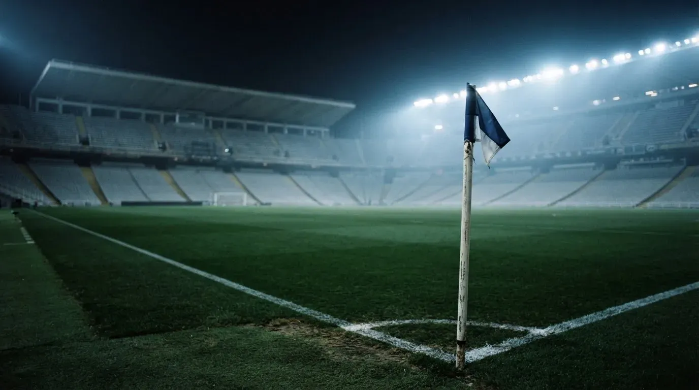 Eine Eckfahne am Rand eines Rasenplatzes, dahinter ein leeres Stadion unter Flutlicht aus der Ferne gesehen.
