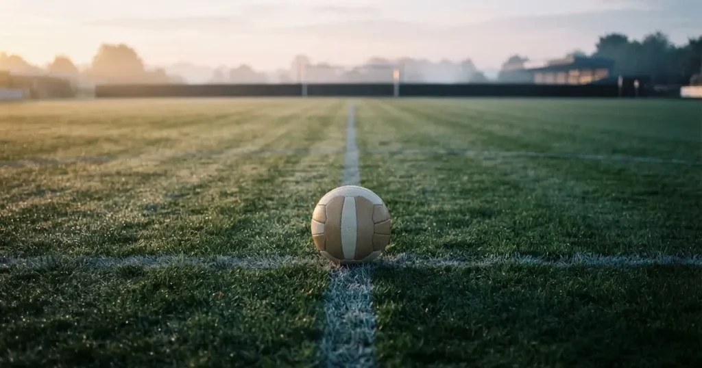 Ein Fußball liegt auf dem Anstoßpunkt eines leeren Rasenplatzes in der Morgendämmerung, Tau glitzert auf dem Gras.