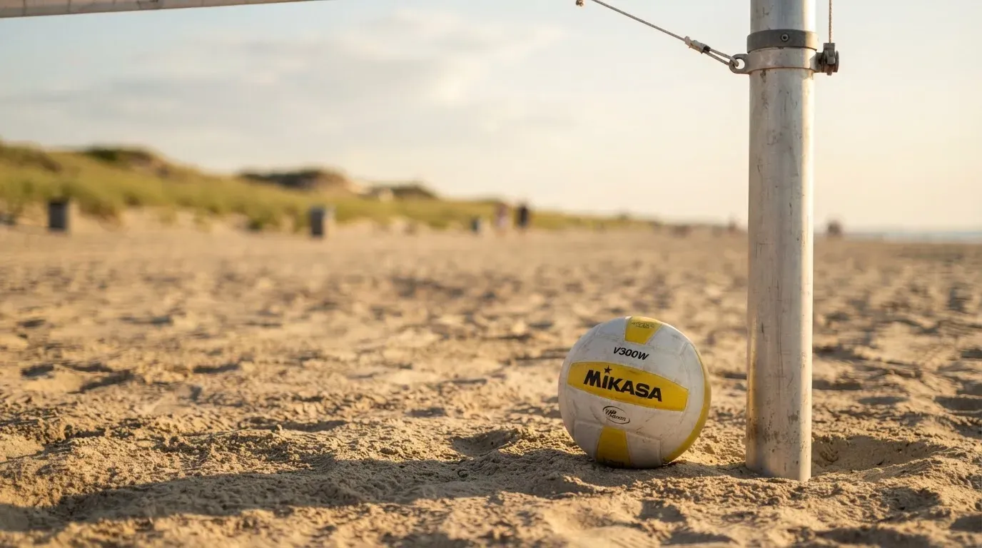 Ein Volleyball liegt auf Sand neben einem Netzpfosten, Strandlicht am späten Nachmittag, im Hintergrund unscharfe Dünen.