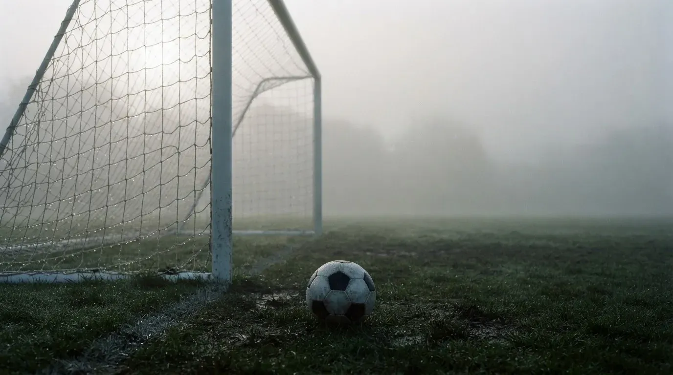 Ein Fußballtor im Nebel eines frühen Morgens, das Netz hängt feucht, auf dem Rasen davor liegt ein einzelner Ball.
