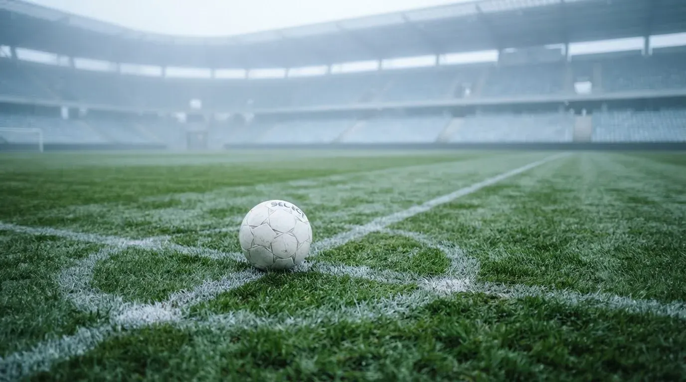Ein Fußball auf dem Mittelpunkt eines Rasenplatzes, früher Morgen, Tau auf dem Gras, im Hintergrund leere Tribünen.