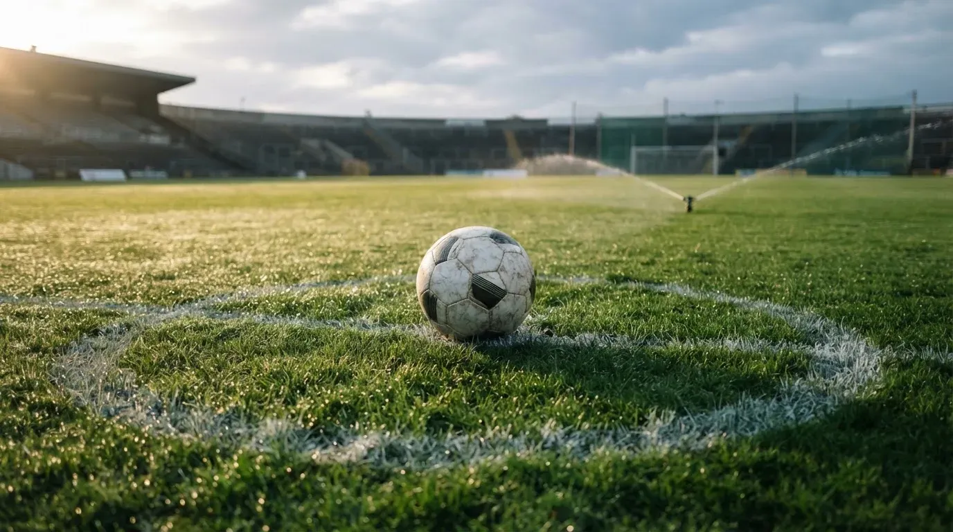 Ein Fußball auf dem Mittelpunkt eines Rasenplatzes, früher Morgen, Tau auf dem Gras, im Hintergrund leere Tribünen.