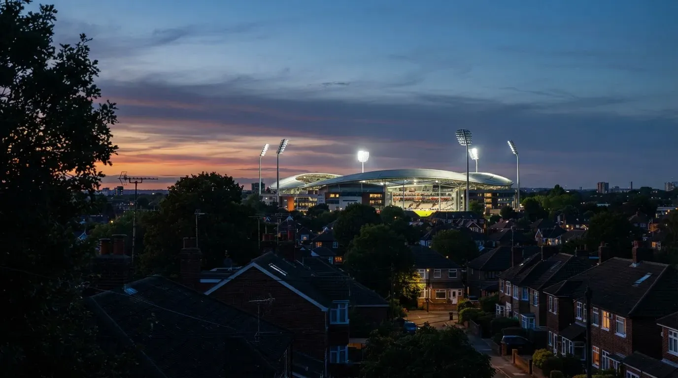Ein Flutlichtstadion bei Dämmerung aus weiter Ferne gesehen, die Lichtmasten leuchten über Dächern, Bäumen und einer ruhigen Stadtrandlandschaft.