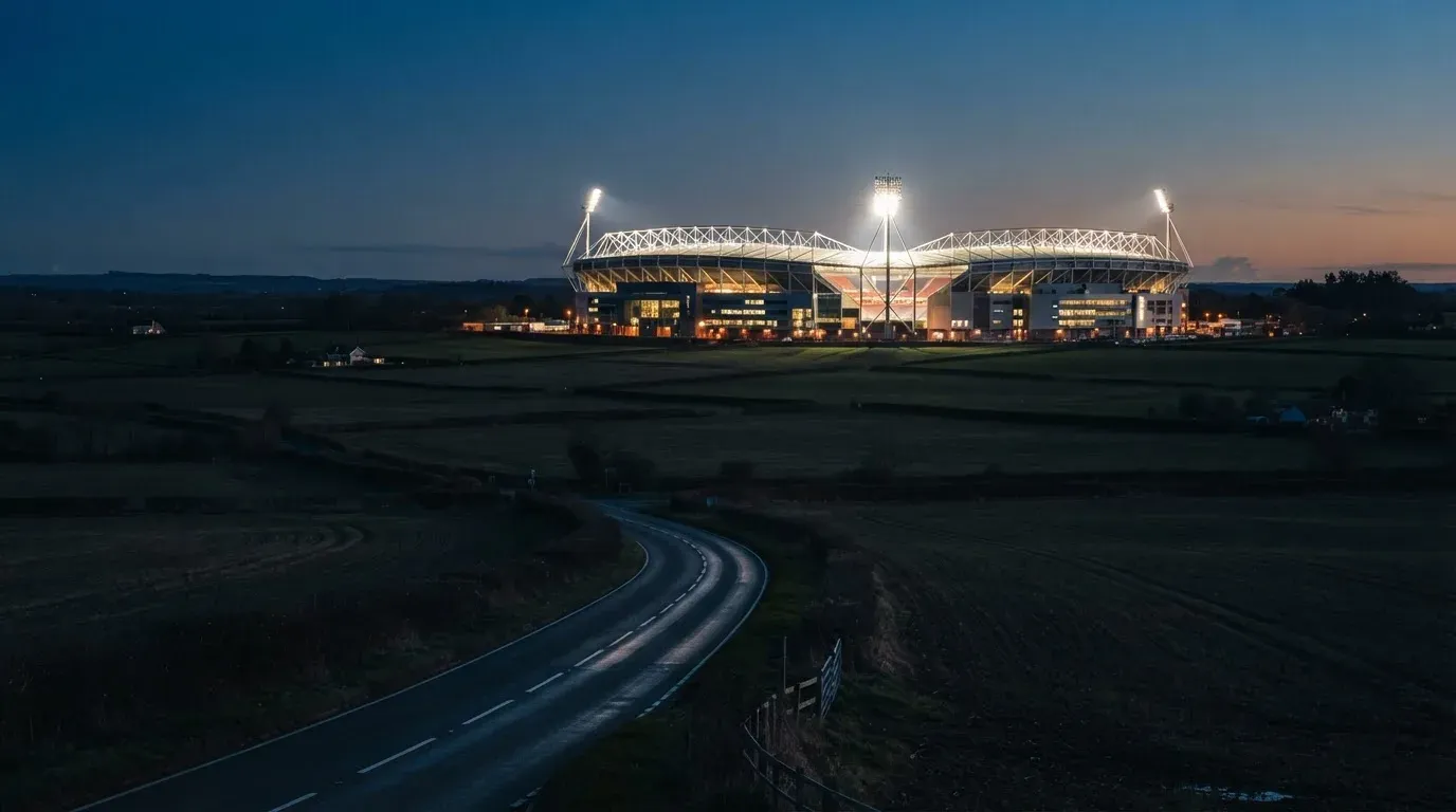 Ein Stadion unter Flutlicht aus der Ferne gesehen, davor eine dunkle Landschaft mit Feldern und einer Landstraße.