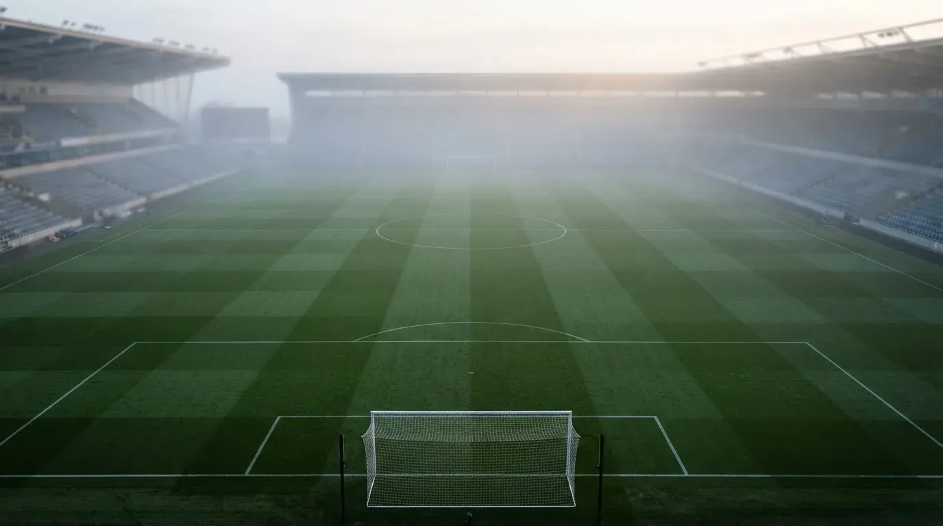 Ein Fußballfeld im Morgennebel aus erhöhter Position gesehen, Mittellinie und Tore sind sichtbar, keine Spieler auf dem Rasen.