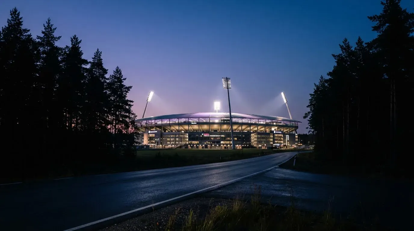 Ein Flutlichtstadion in der Dämmerung aus weiter Ferne, davor ein Wald und eine leere Straße.