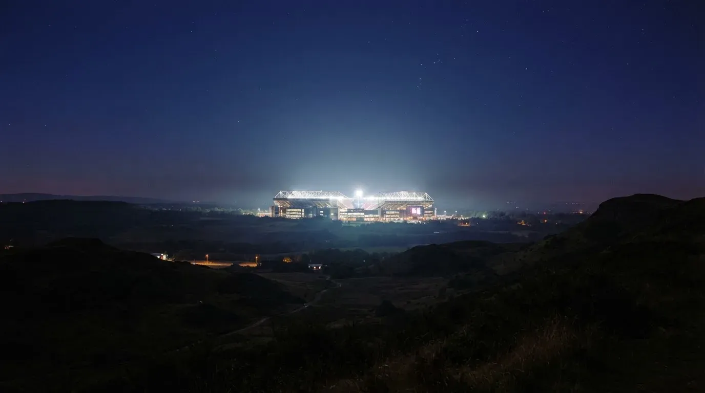 Ein Stadion unter Flutlicht aus großer Entfernung gesehen, darüber ein klarer Nachthimmel und davor dunkle Hügel.