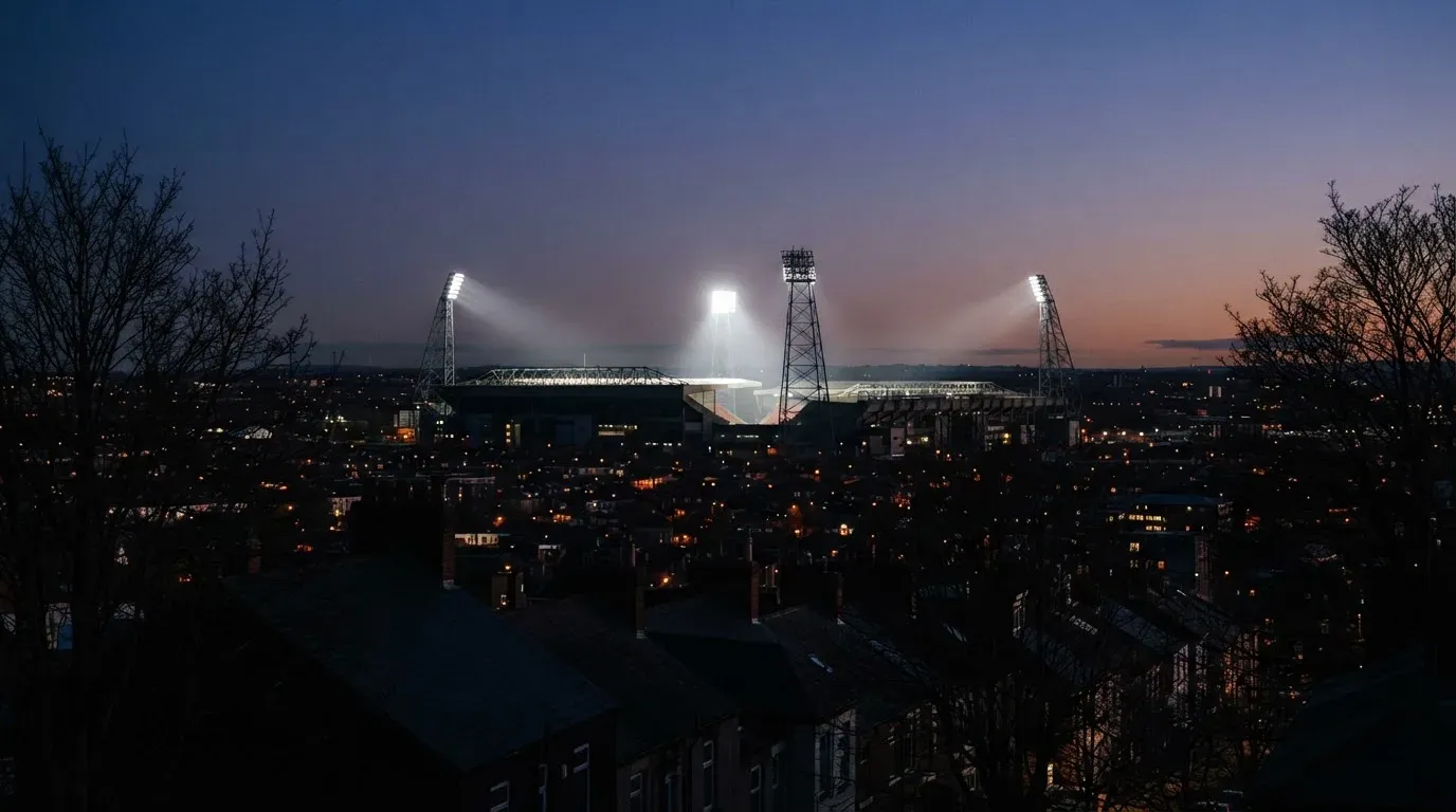 Ein Flutlichtstadion bei Dämmerung aus weiter Ferne gesehen, die Lichtmasten leuchten über Dächern und Bäumen.