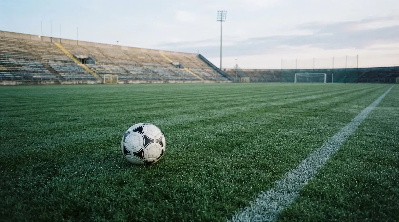Ein Fußball liegt auf einem Rasenplatz im Morgenlicht, im Hintergrund eine ruhige Tribüne ohne Zuschauer.