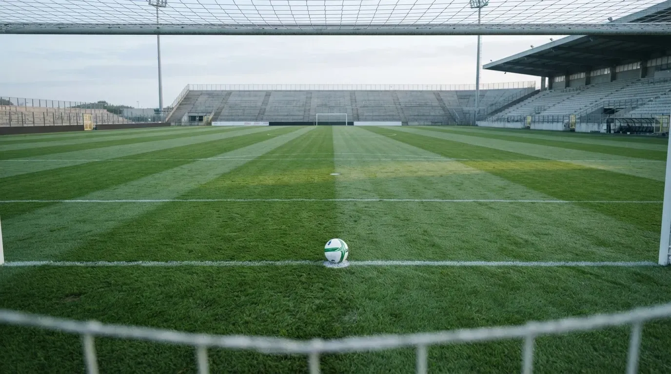 Ein Fußball liegt auf dem Elfmeterpunkt eines leeren Rasenplatzes im Morgenlicht, dahinter eine ruhige Tribüne ohne Zuschauer.