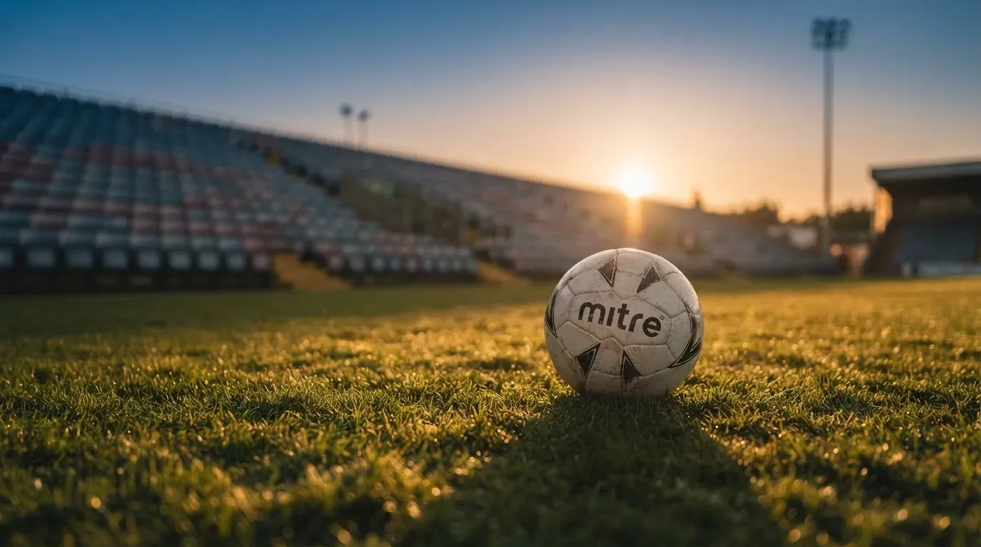 Ein Fußball liegt auf einem Rasen im Licht der tief stehenden Sonne, dahinter leere Sitzreihen.