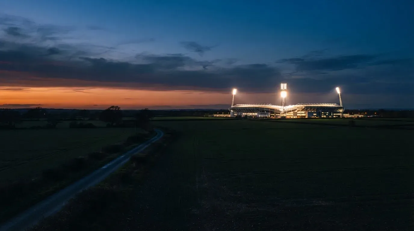 Ein Stadion unter Flutlicht aus der Ferne gesehen, davor eine dunkle Landschaft mit Feldern und einer Landstraße.