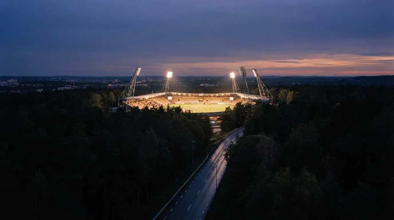 Ein Flutlichtstadion in der Dämmerung aus weiter Ferne, davor ein Wald und eine leere Straße.