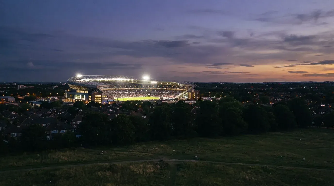 Ein Flutlichtstadion bei Dämmerung aus weiter Ferne gesehen, die Lichtmasten leuchten über Dächern und Bäumen.