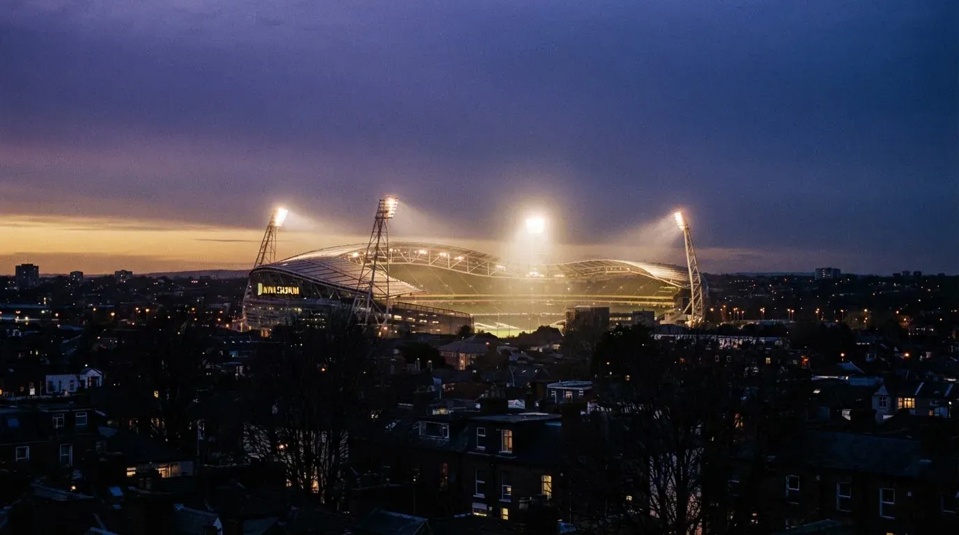 Ein Flutlichtstadion bei Dämmerung aus weiter Ferne gesehen, die Lichtmasten leuchten über Dächern und Bäumen.